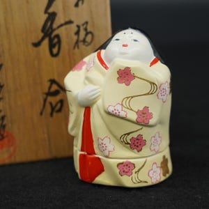 以下が含まれることがあります： 黒髪の人物の陶器製フィギュア。ピンクと白の桜模様が描かれたクリーム色の着物を着ており、鮮やかな赤色の縦のアクセントが入っている。黒い日本語の書が書かれた木箱の前に置かれている。