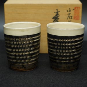 以下が含まれることがあります： 黒と白のダイヤモンド模様のセラミックカップ2個。上部の縁は白く、下部は濃い茶色です。カップは円筒形で、暗い面に置かれています。背景には木箱があります。