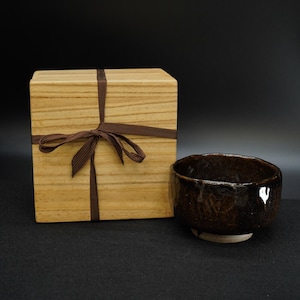 May include: A dark brown ceramic tea bowl with a textured surface and a white base sits next to a wooden box tied with a brown ribbon. The box has a light wood grain pattern.