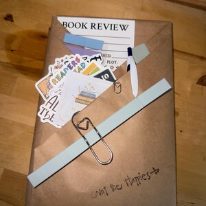 May include: A wrapped package with a "Book Review" label, adorned with stickers, sticky notes, and a heart-shaped paper clip. The package is sealed with a light blue tape and has the handwritten text "Count the Happies".