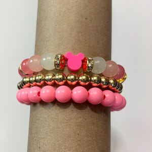 May include: Stack of three beaded bracelets in shades of pink and gold. The top bracelet features pink and off-white beads with a gold accent and a pink Mickey Mouse charm. The middle bracelet has gold beads, and the bottom bracelet has pink beads.