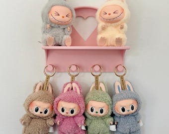 May include: A pink shelf displays six plush bunny figures. Two are seated, wearing hooded costumes with smiling faces. Below, four keychains in brown, pink, green, and blue feature similar bunny characters with stern expressions. The text "@blindboxboutique" is visible.