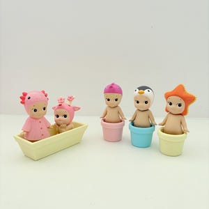 Puede incluir: Una colección de pequeñas y caprichosas figuras. Dos figuras están en un bote amarillo y tres en macetas de colores. Las figuras tienen varios tocados, incluyendo un dinosaurio rosa, un ciervo, una magdalena rosa, un pingüino y una estrella naranja.
