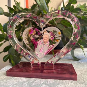 Peut inclure: Un cadre photo en résine en forme de cœur avec une photo d'une femme entourée de fleurs. La résine transparente est incrustée de pétales de fleurs roses et mauves. Le cadre repose sur une base rectangulaire de couleur bordeaux.