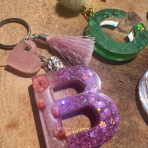 Puede incluir: Una colección de llaveros hechos a mano con letras y dijes de resina. La letra "B" es de un morado vibrante con purpurina y flores secas. Otros llaveros incluyen un corazón rosa, la letra "C" verde y un pompón verde esponjoso.