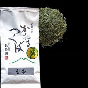 Op de afbeelding: Een witte verpakking Japanse thee met Japanse tekst en een hoop losse groene theebladeren. De verpakking heeft zwarte en groene Japanse karakters en een groene cirkel met Japanse tekst. De theebladeren zijn heldergroen.