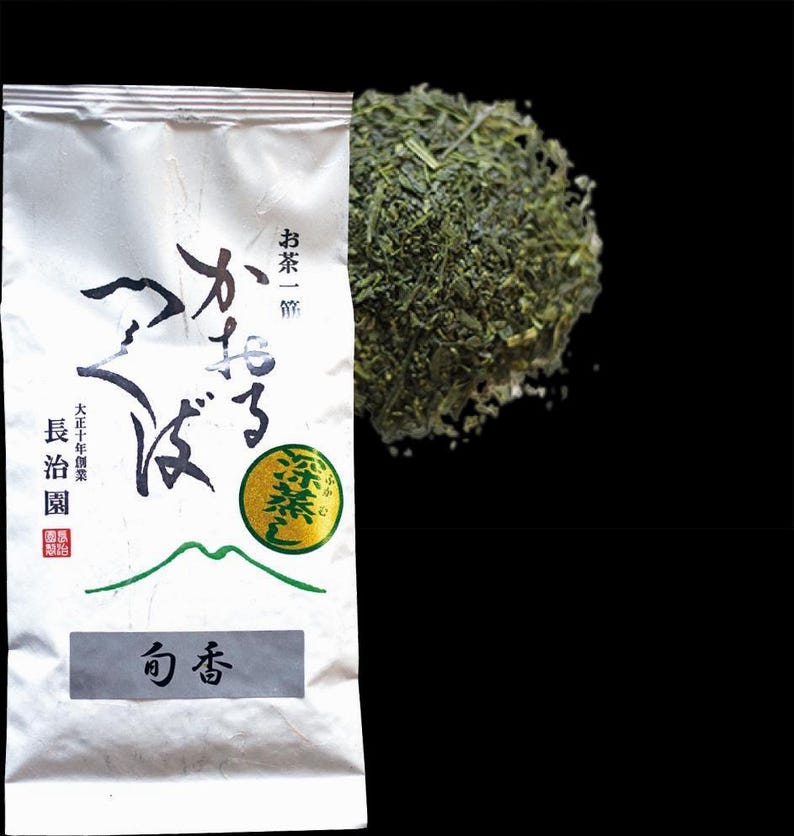 Op de afbeelding: Een witte verpakking Japanse thee met Japanse tekst en een hoop losse groene theebladeren. De verpakking heeft zwarte en groene Japanse karakters en een groene cirkel met Japanse tekst. De theebladeren zijn heldergroen.