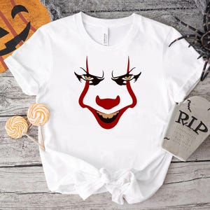 Può includere: T-shirt bianca con grafica di un volto da clown con dettagli rossi e un'espressione sinistra. Il design include naso, bocca e segni sul viso rossi. Tema Halloween con zucca e ragno.