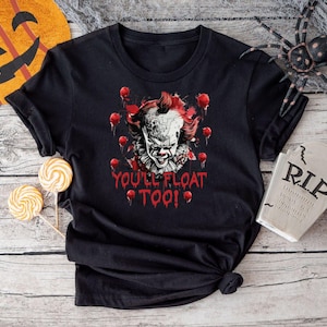 May include: Black t-shirt featuring a scary clown graphic with the text "YOU'LL FLOAT TOO!". The design includes red balloons and dripping blood, perfect for Halloween. The shirt is displayed with Halloween decorations.