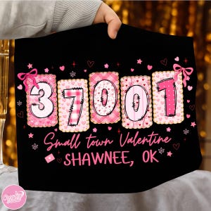 May include: Black t-shirt with the numbers "37001" in pink and white, decorated with hearts and bows. The text "Small town Valentine SHAWNEE, OK" is printed below the numbers. The shirt is held up by a person.