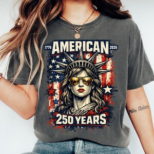 May include: Dark grey t-shirt with a graphic of the Statue of Liberty wearing sunglasses. The design includes the text "AMERICAN 250 YEARS" and the dates "1776" and "2026", with the colours of the American flag.