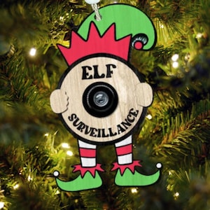 以下が含まれることがあります： 「ELF SURVEILLANCE」の文字が入った、エルフの形をした木製クリスマスオーナメント。 オーナメントの中央にはカメラレンズがあり、赤、緑、白のアクセントが施されています。 クリスマスツリーに飾られています。