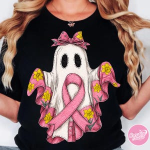 以下が含まれることがあります： ピンクのリボン、リボン、花のディテールを取り入れた、白い幽霊のデザインの黒いTシャツ。幽霊は大きな楕円形の目を持ち、ピンクのリボンで飾られ、袖とリボンには黄色の花が咲いています。乳がん啓発のデザインです。
