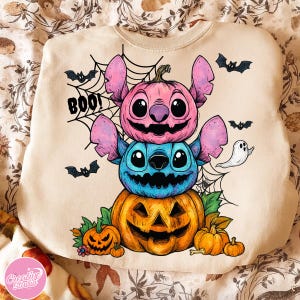 May include: Beige sweatshirt featuring a Halloween-themed design. The design includes a stack of three carved pumpkins, each resembling a character with pink and blue ears. The word "BOO!" is written above the pumpkins, with bats and spiderwebs.