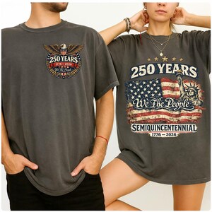 May include: Two grey t-shirts with patriotic designs. One has an eagle emblem with "250 YEARS 1776-2026". The other shows an American flag, the Statue of Liberty, and the words "250 YEARS We The People SEMIQUICENTENNIAL 1776-2026".