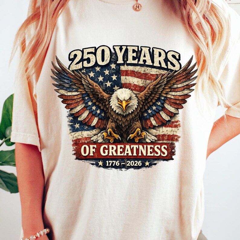 May include: A cream-coloured t-shirt with a patriotic design. It features a bald eagle with outstretched wings, set against a distressed American flag. Text reads "250 YEARS OF GREATNESS 1776-2026" in bold lettering.