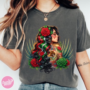 May include: Dark grey t-shirt featuring a detailed illustration of a woman with long, dark hair adorned with red roses and the colors of the Mexican flag. The woman has tattoos and is surrounded by green and gold accents.