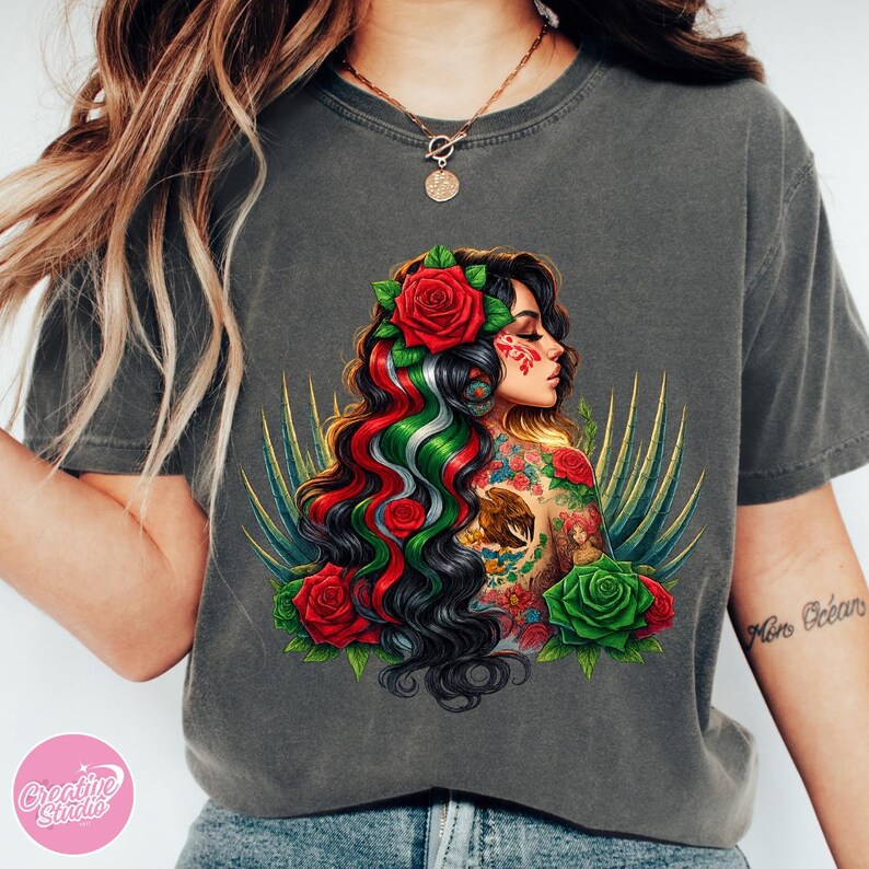 May include: Dark grey t-shirt featuring a detailed illustration of a woman with long, dark hair adorned with red roses and the colors of the Mexican flag. The woman has tattoos and is surrounded by green and gold accents.