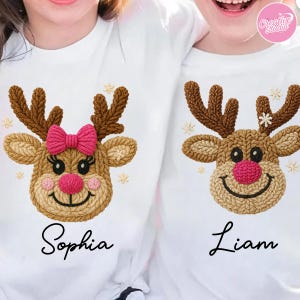 Puede incluir: Camisetas blancas con un diseño de reno tejido. Un reno tiene un lazo rosa, el otro un copo de nieve. Los nombres "Sophia" y "Liam" están escritos debajo. Los renos tienen cuernos marrones, narices rosas y caras sonrientes.