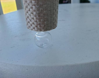 Op de afbeelding: Een miniatuurlamp met een geweven jute lampenkap en een heldere glazen voet. De cilindrische kap heeft een witte binnenkant. De lamp is ontworpen om op een tafel of ander oppervlak te staan.