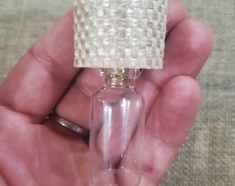 Op de afbeelding: Een miniatuurlamp met een heldere glazen voet en een geweven, crèmekleurige lampenkap. De lamp wordt in een hand gehouden, wat de kleine afmetingen en het gedetailleerde ontwerp benadrukt. De lampenkap heeft een getextureerde afwerking.