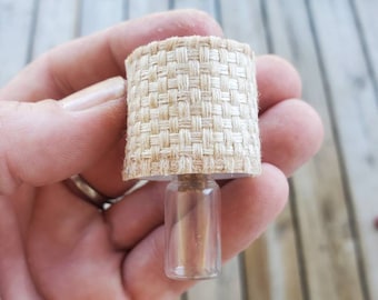 Op de afbeelding: Miniaturlamp met een geweven beige lampenkap en een heldere glazen voet. De lamp is klein, ontworpen voor een poppenhuis of een vergelijkbare setting. De lampenkap heeft een getextureerd uiterlijk.