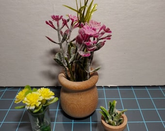 May include: Three miniature potted plants. A small wooden pot holds pink flowers and green foliage. A clear glass holds yellow flowers. A small wooden pot holds a green succulent.