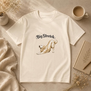 Może przedstawiać: Kremowy t-shirt z krótkim rękawem z brązową ilustracją liniową przedstawiającą psa rasy golden retriever w pozycji rozciągania. Nad psem znajduje się napis "Big Stretch" wydrukowany ciemnobrązową, zakrzywioną czcionką odręczną.
