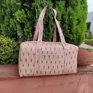 May include: A rectangular quilted duffel bag featuring a beige base with a repeating pattern of small pink floral motifs and dark blue stems. The bag includes two long handles with a pink and white striped design and a matching striped trim.