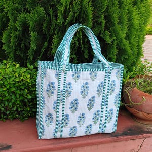 May include: White tote bag with a blue floral pattern. The bag has green and white striped handles and trim. The bag is sitting on a red surface with green foliage in the background.