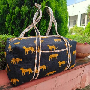 May include: Black duffel bag with a repeating pattern of yellow tigers. The bag has a rectangular shape with a zipper closure and two handles. The handles and trim are striped with white, tan, and light blue. The bag is sitting on a red surface.