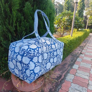 May include: A blue and white floral patterned duffel bag with matching handles. The bag is rectangular and appears to be made of fabric. The bag is sitting on a brown pot, with a green hedge and trees in the background.