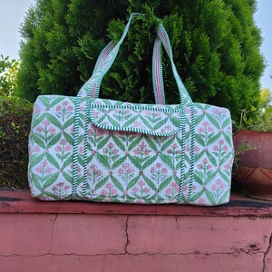 Könnte beinhalten: Eine weiße und grüne Tragetasche mit floralem Muster und rosa Akzenten. Die Tasche hat eine rechteckige Form mit einem Klappenverschluss und zwei Griffen. Die Tasche ist aus einem gewebten Stoff gefertigt.