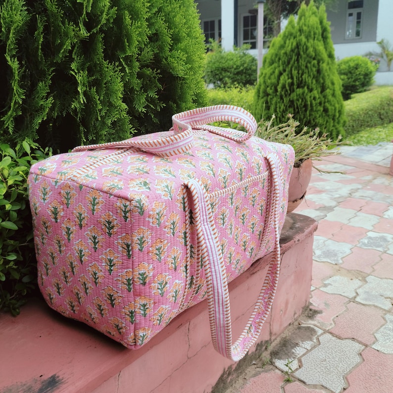 May include: Pink floral patterned travel bag with matching handles. The bag features a quilted design with a repeating pattern of yellow and green flowers. The handles are striped with white and gold. The bag is sitting on a pink surface.