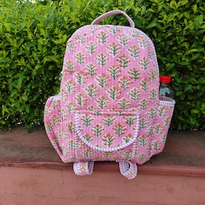 May include: A quilted backpack in a light pink fabric featuring a repeating pattern of orange and green floral motifs. The bag includes a top handle, a front pocket with a flap closure, and side pockets, one of which holds a clear water bottle.
