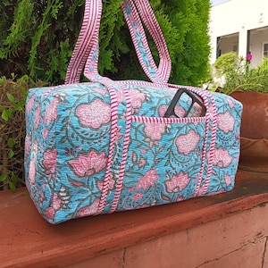 May include: A rectangular, quilted tote bag with a floral pattern in pink and blue. The bag has a front pocket and striped pink and white handles. A pair of black glasses is in the front pocket.