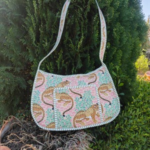 Puede incluir: Bolso de hombro rosa con un estampado repetido de guepardos y follaje verde. El bolso tiene un ribete blanco y verde claro y una correa a juego. El bolso está hecho de tela y está diseñado para llevar objetos.