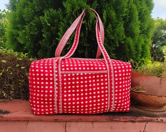 Puede incluir: Un bolso tote a cuadros rojos y blancos con asas a juego. El bolso tiene forma rectangular y cierre de cremallera. Las asas son de un rojo más claro con un diseño blanco. El bolso está sobre una superficie roja con follaje verde al fondo.
