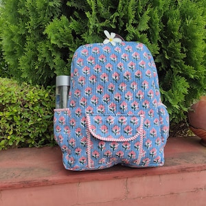 Puede incluir: Una mochila azul acolchada con un patrón floral repetitivo en rosa y verde. La bolsa incluye un bolsillo frontal con solapa, bolsillos laterales y una botella de agua transparente. Una flor blanca descansa sobre el asa superior.