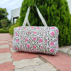 Puede incluir: Un bolso tote blanco con un estampado floral rosa y hojas verdes. El bolso tiene dos asas y forma rectangular. El bolso está hecho de tela acolchada.