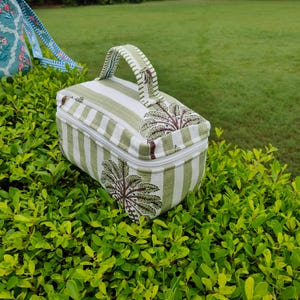 Puede incluir: Una bolsa rectangular de tela a rayas verdes y blancas con asa. La bolsa tiene cierre de cremallera y un diseño decorativo de árbol en las esquinas. La bolsa está sobre un lecho de hojas verdes.