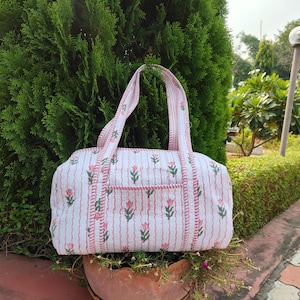 May include: A white and pink floral patterned tote bag with matching handles. The bag features a vertical striped design with pink flowers and green leaves. The bag is sitting on a brown pot.