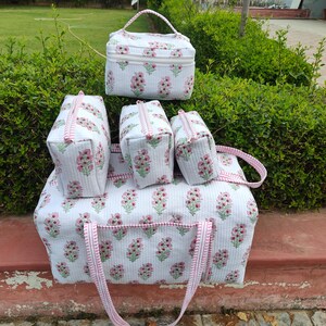 May include: A set of white quilted travel bags and a cosmetic bag, adorned with a pink floral pattern. The set includes a large tote bag, a cosmetic bag with a handle, and three smaller zippered bags. The bags have pink striped trim.