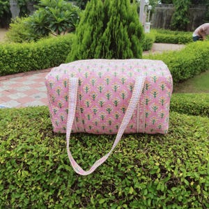May include: A pink quilted tote bag with a floral pattern in yellow and green. The bag has two white and pink striped handles. The bag is sitting on a green bush.