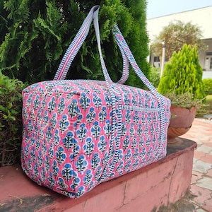 Peut inclure: Un sac fourre-tout à motifs floraux roses et bleus avec des sangles assorties. Le sac a une forme rectangulaire et est fait de tissu matelassé. Les sangles sont blanches avec des rayures roses et bleues. Le sac est posé sur une surface rouge.