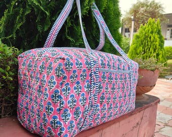 May include: A pink and blue floral patterned tote bag with matching straps. The bag has a rectangular shape and is made of quilted fabric. The straps are white with pink and blue stripes. The bag is sitting on a red surface.