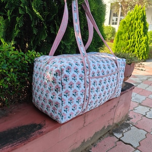 Puede incluir: Una bolsa de lona con estampado floral blanco y rosa con asas a rayas rosas. La bolsa es rectangular y parece estar hecha de tela acolchada. La bolsa está sobre una superficie roja con follaje verde en el fondo.