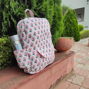 Puede incluir: Una mochila de tela acolchada con un patrón floral repetitivo en rosa y verde sobre un fondo blanco. La mochila incluye un asa superior, un bolsillo frontal con solapa y un bolsillo lateral que contiene una botella de agua transparente con tapa plateada.