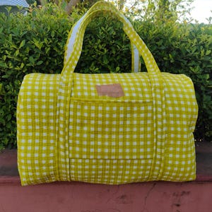 May include: A large, rectangular duffel bag in a yellow and white checkered pattern. The bag has a front pocket and two handles. The handles are yellow with a white stripe down the middle. A small, brown leather patch is sewn on the front.