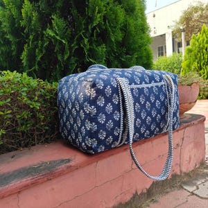 May include: A navy blue and white patterned duffel bag with a floral design. The bag has two handles and a long shoulder strap. The bag is sitting on a red brick surface, with green foliage in the background.